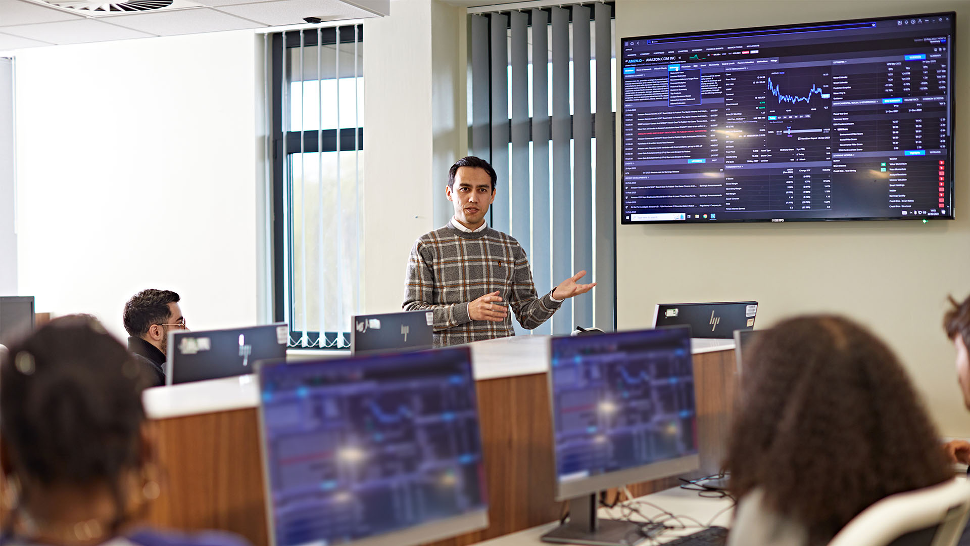 Trading room with students working on laptops and teacher in front of a large trading screen