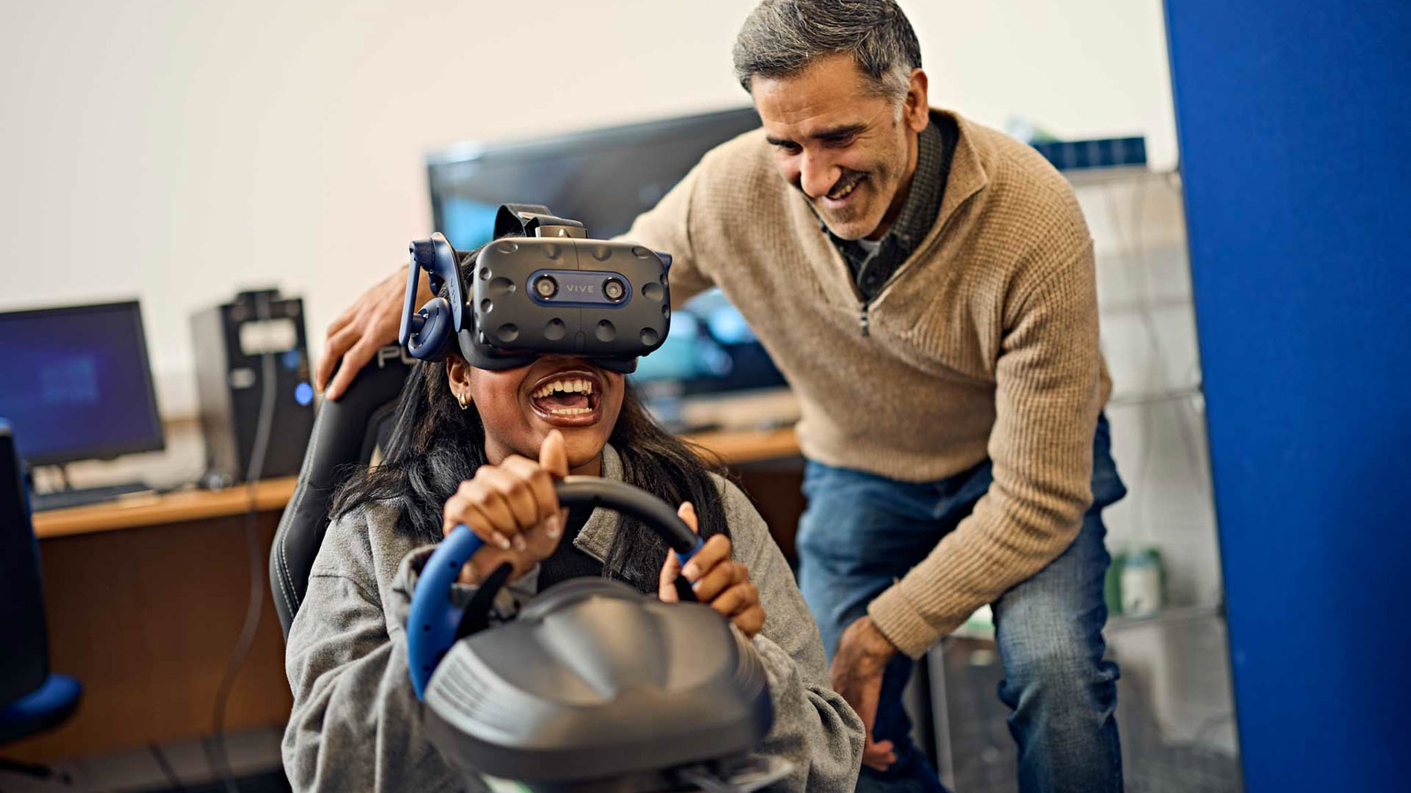 A psychology student and lecturer conducting a simulated driving experiment using virtual reality equipment. The student drives a virtual vehicle in a simulated environment using a driving simulator and VR headset while the lecturer looks on.